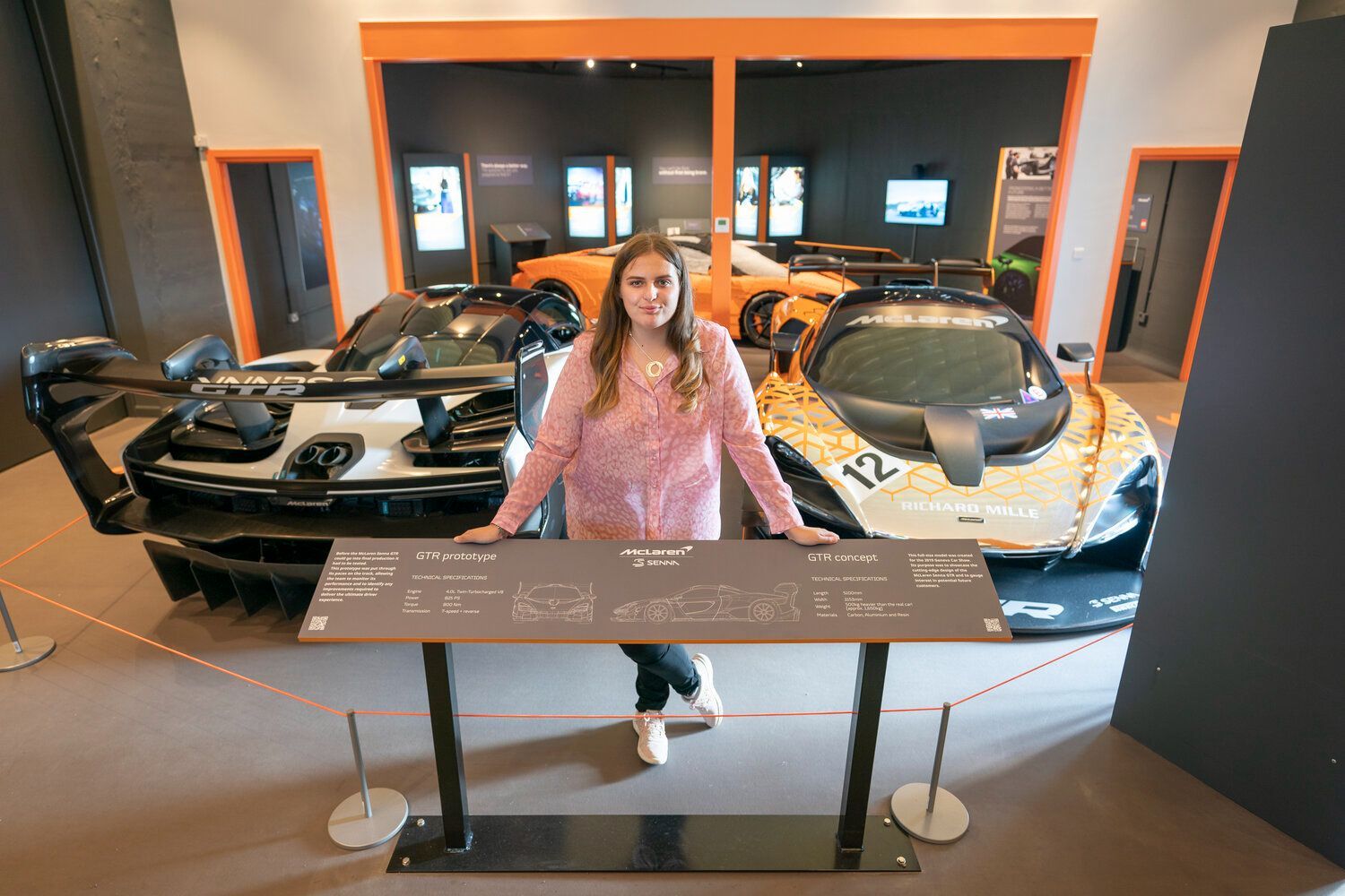 Yound woman in pink top stand inside the exhibition, with the white McLaren Senna GTR Prototype and Orange and silver Concept cars behind