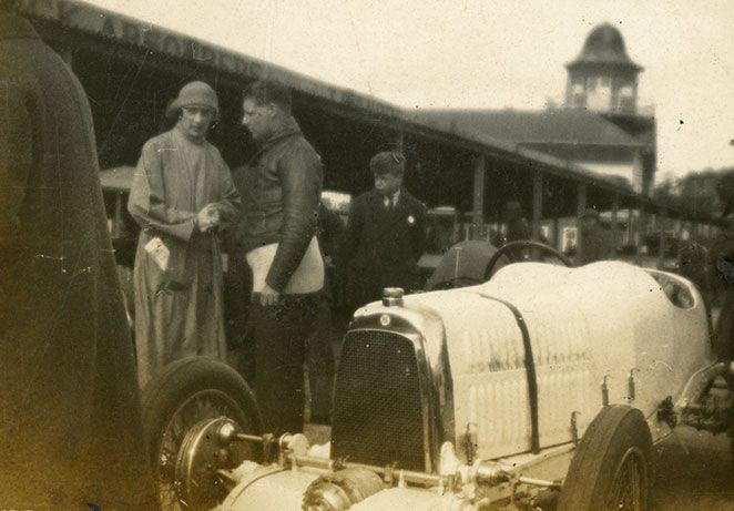 Vintage race car in the foreground, behind a man and a women in 20s clothing chat, Brooklands race bays and Clubhouse visible behind