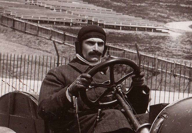 Man in victorian style driving gear holding the very large wheel of a vintage car.