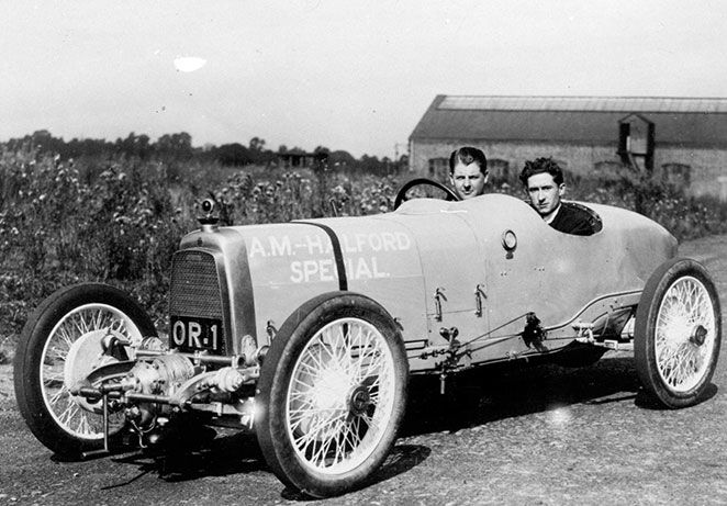 Two men sat in a vinatge race car, fields and a barn in the background