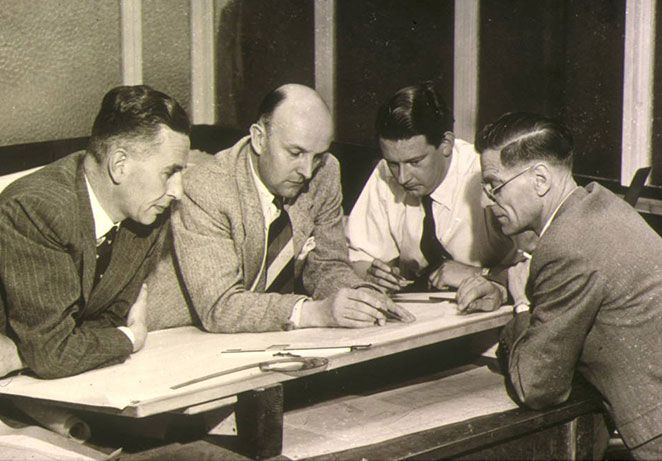 four men in suites looking down at a design on a desk