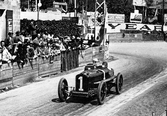 vintage race car with a white 1 painted on front, driven around a race track with crowds in the background