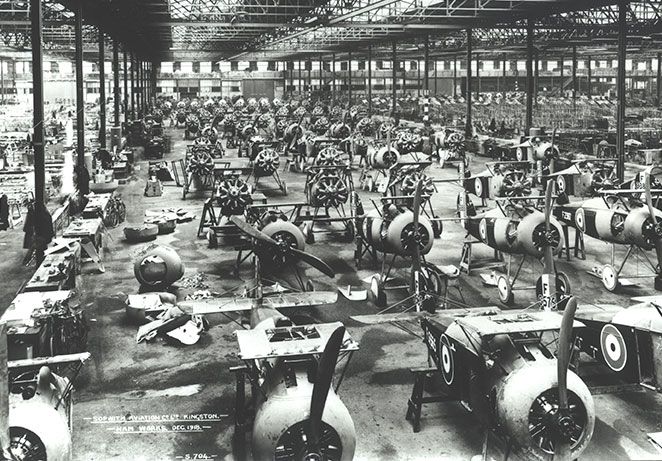 Busy factory scene, long lines of partial built biplanes inside a large open hangar.
