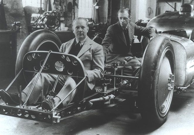 Two men in suits, one sat in in the frame of a race car, with the second crouched behind