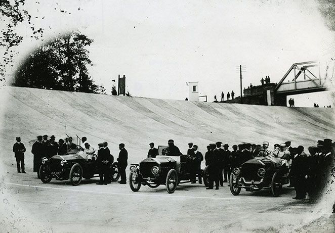 Three vintage car on the banked race track surrounded by people.
